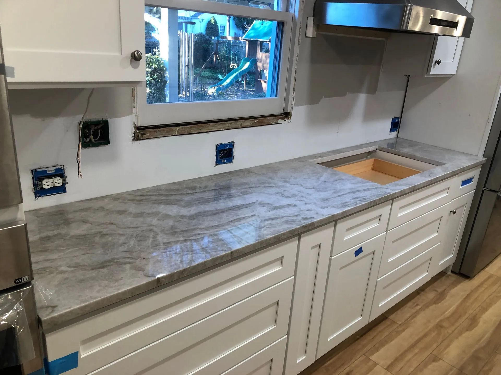 Kitchen countertop installation: White cabinets, gray granite, under-window sink space, unfinished wall with electrical boxes.