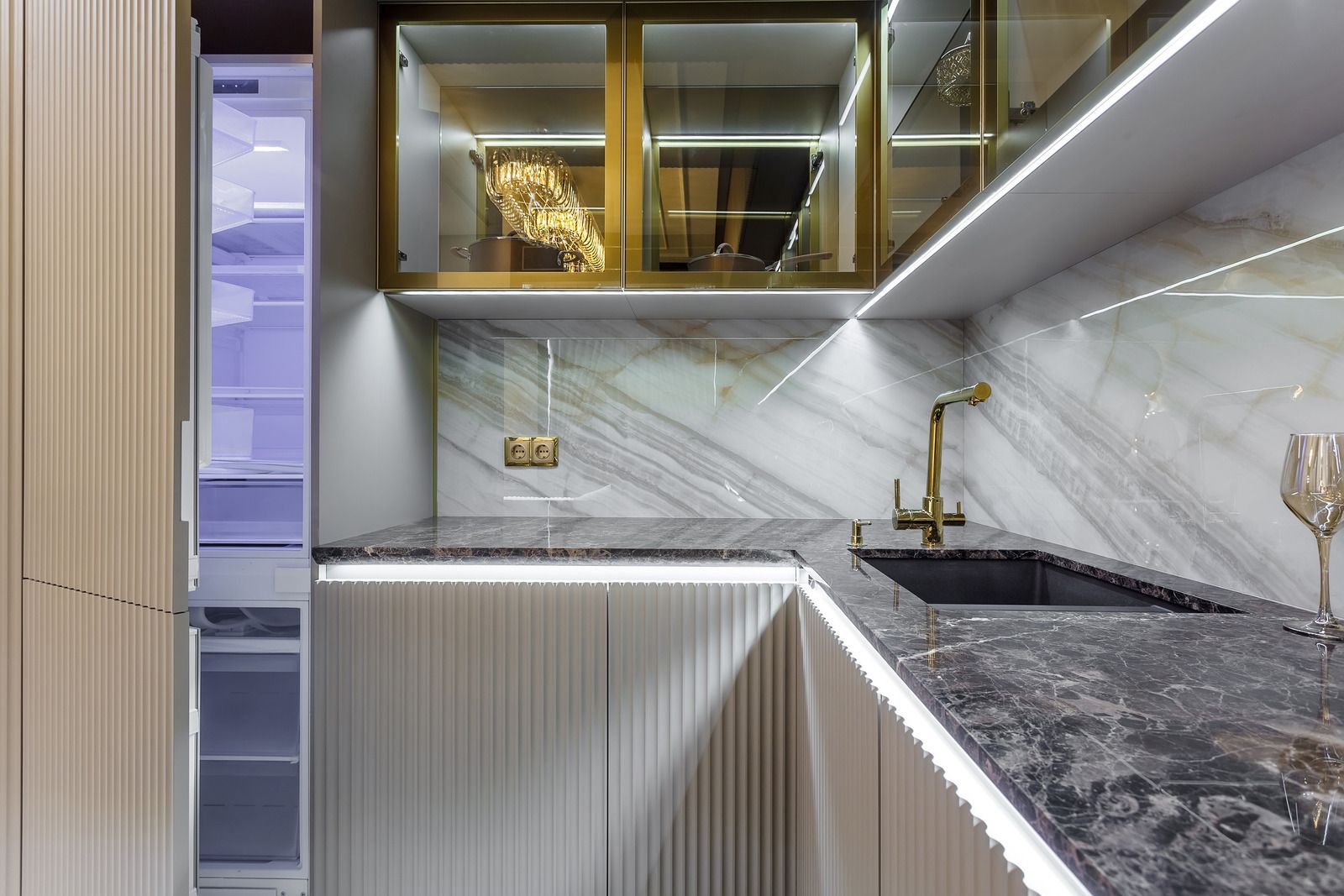 Modern kitchen with marble countertops, gold-framed glass cabinets, and a sink. Built-in refrigerator and led lighting.