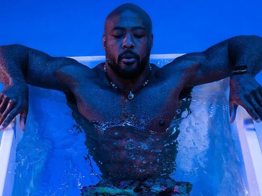 A shirtless man is laying in a bathtub with his arms outstretched.