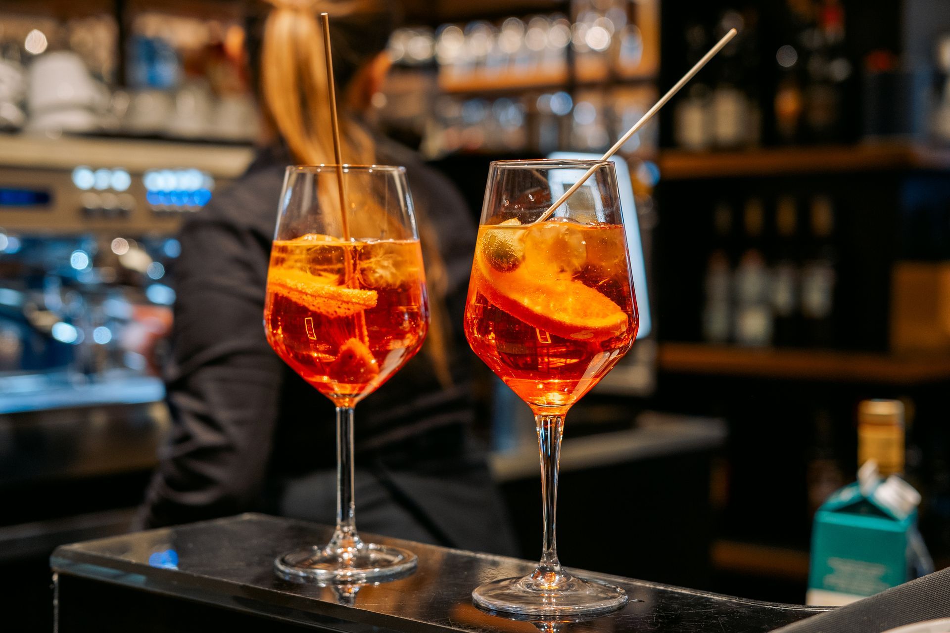 Due cocktail Aperol Spritz con fette d'arancia e stirrer su un bancone; il barista sullo sfondo.