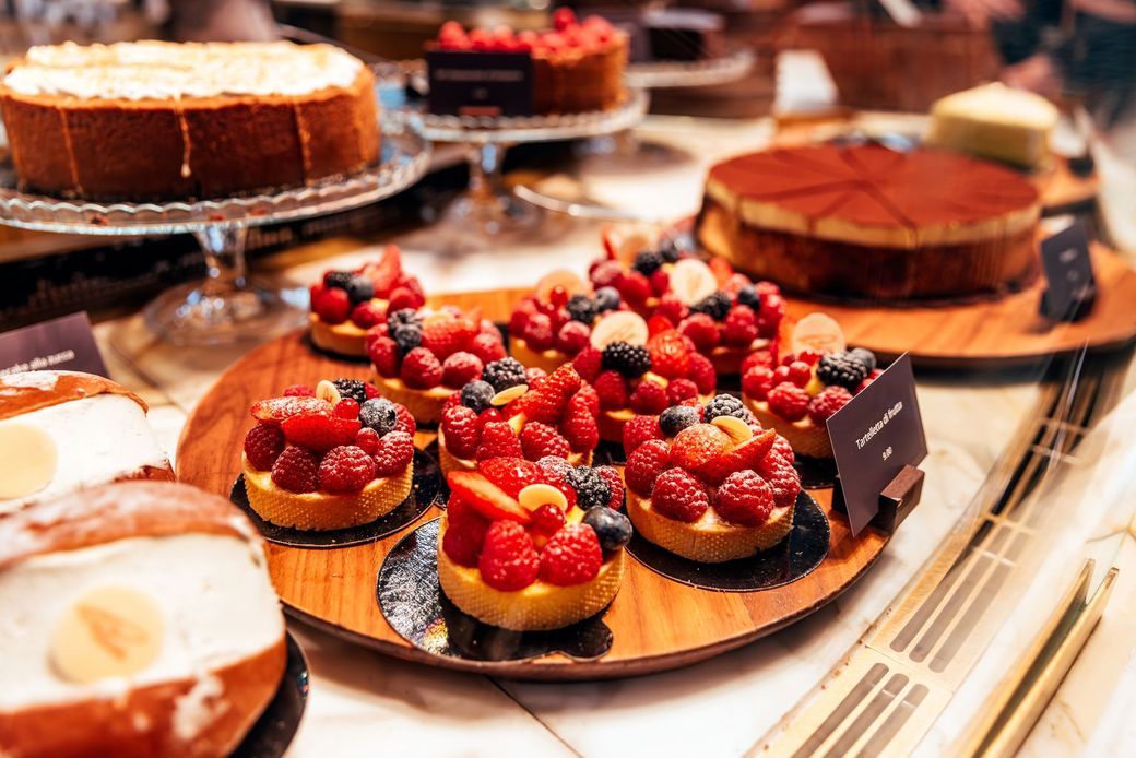 Dolci in esposizione: crostate di frutta, cheesecake e altre torte. Vassoi di legno e alzate di vetro in una panetteria.