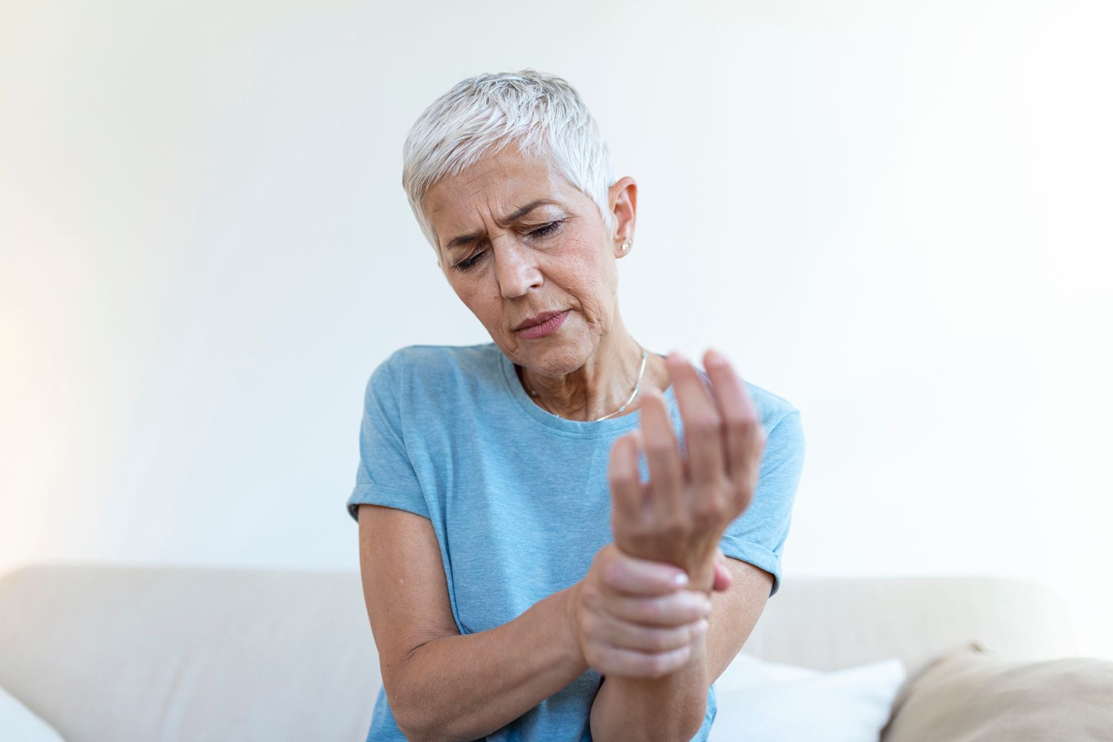 Mujer sujetándose la muñeca, con aspecto de dolor. Está en un lugar cerrado, con camisa azul, posiblemente con dolor.
