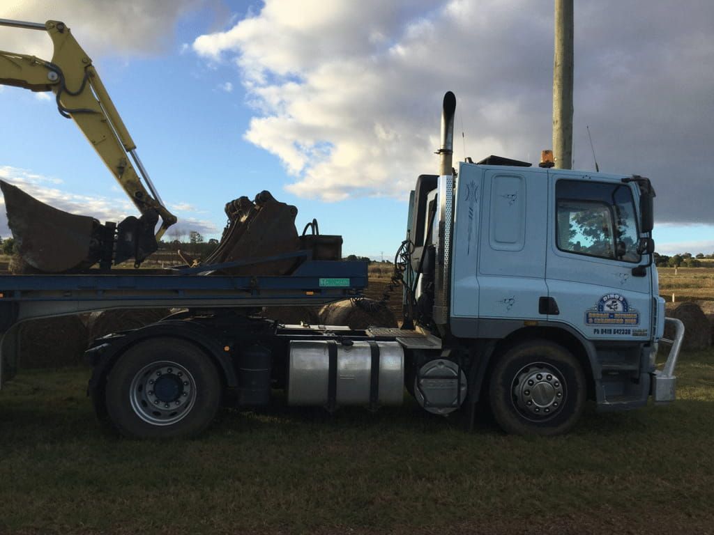 A Truck With a Yellow Excavator on the Back is Parked in a Field — Dino's Bobcat–Bitumen & Excavator Hire in Cushnie, QLD