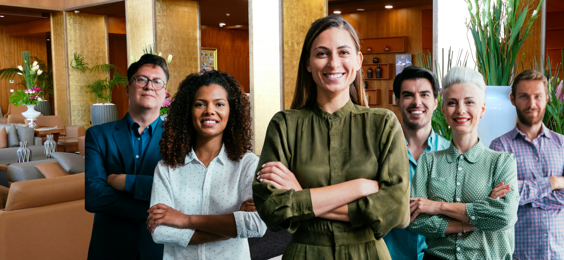 Group of six coworkers standing in a modern office lobby, smiling with arms crossed