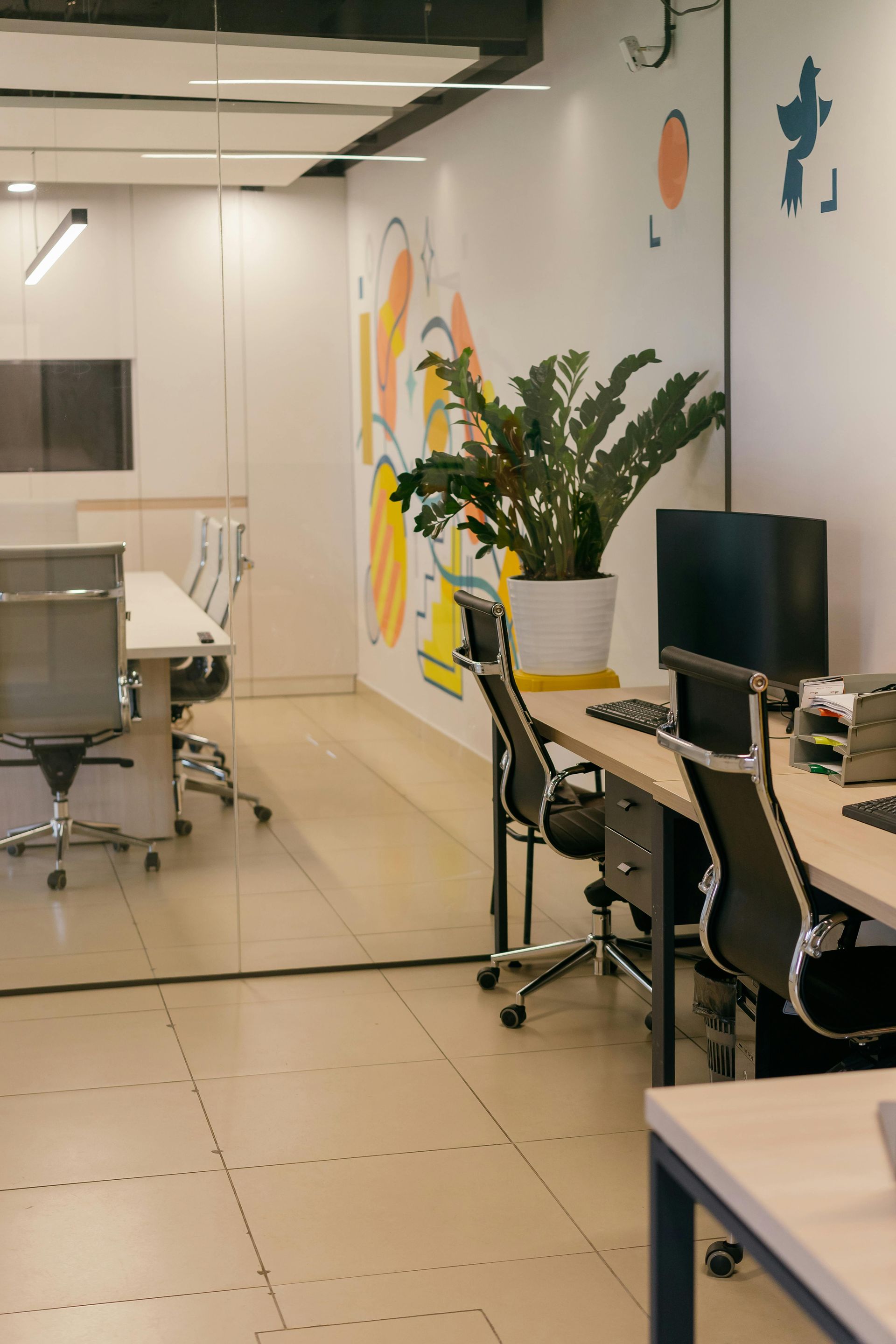 Modern office corridor with desks, computer monitors, chairs, and a large potted plant