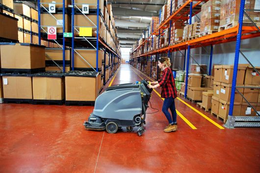 Worker pushing a floor scrubber in a warehouse aisle between tall shelving racks.