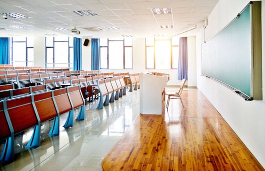 Empty classroom with rows of desks, a podium, and a large chalkboard beside sunlit windows.