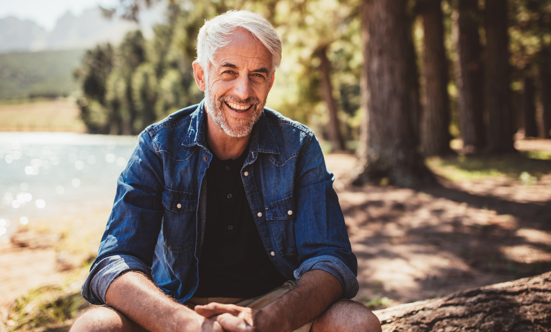 A man is sitting on a rock by a lake with his legs crossed and smiling.