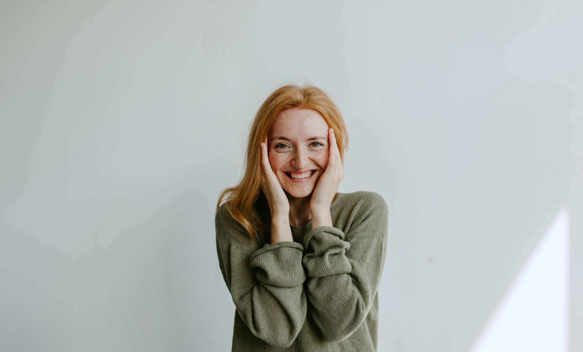 A woman in a green sweater is smiling and covering her face with her hands.