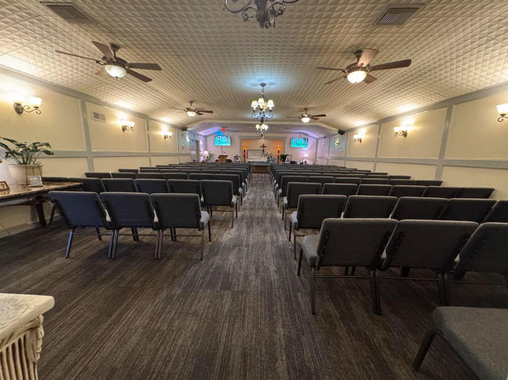 Rows of empty chairs face a stage, in a chapel with ornate ceiling and lights.