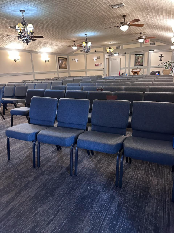 Rows of blue upholstered chairs in a church. Ceiling fans and decorative wall lights.