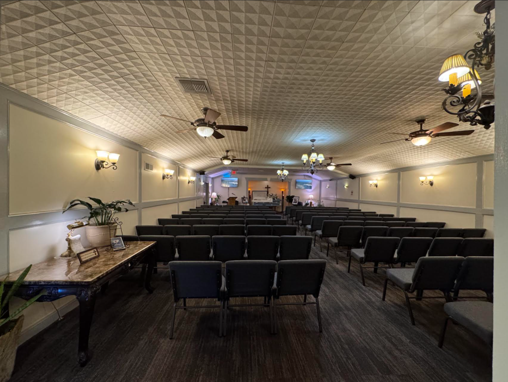 Rows of black chairs face a stage in a chapel with ornate ceiling and wall sconces.