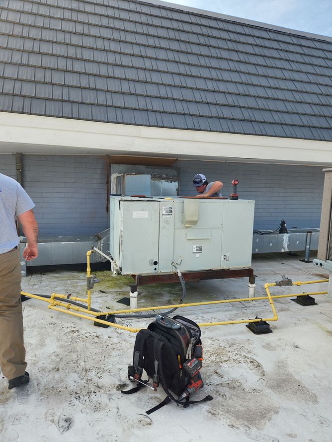 Person working on HVAC unit on a rooftop. Another person stands nearby. Gray roof, backpack on ground.