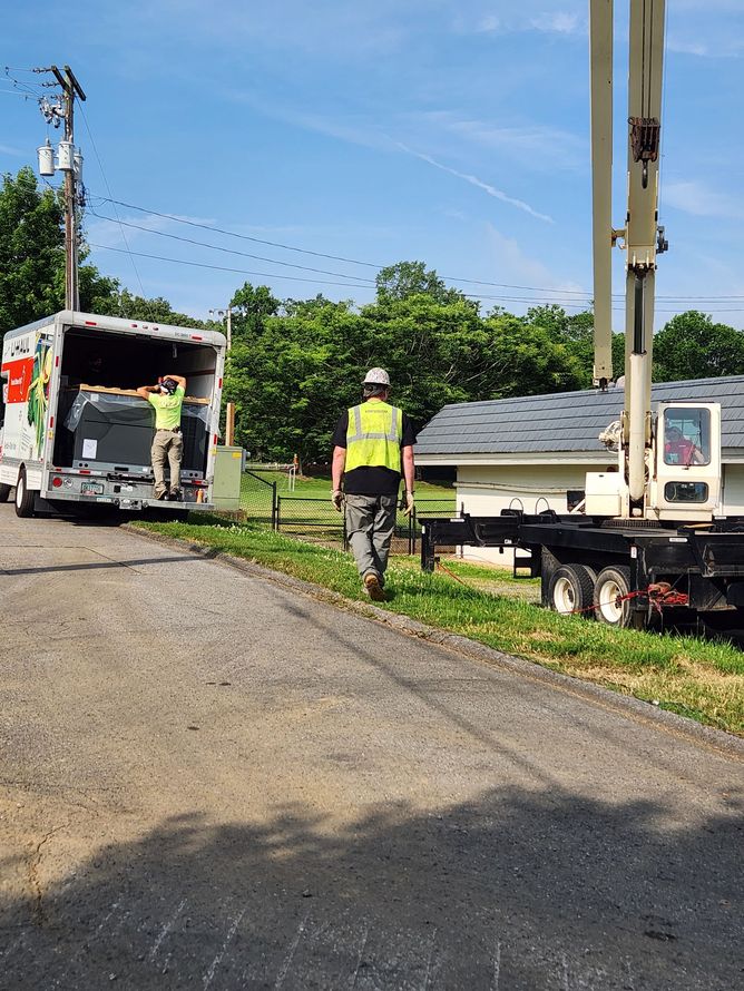 Two workers unloading from a truck with a crane, on a gravel road near a building, under a blue sky.