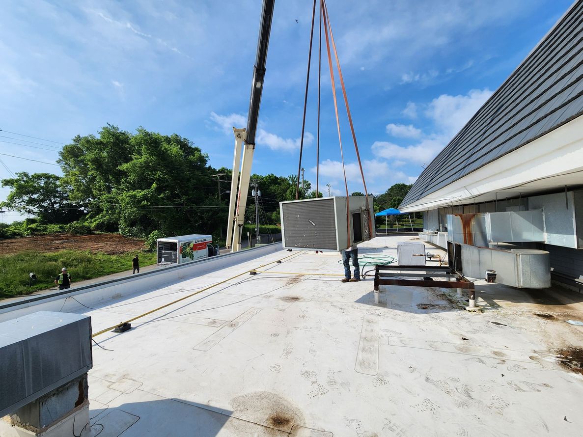 Crane lifting a rectangular unit onto a flat roof. Workers stand nearby. Blue sky, green trees in background.