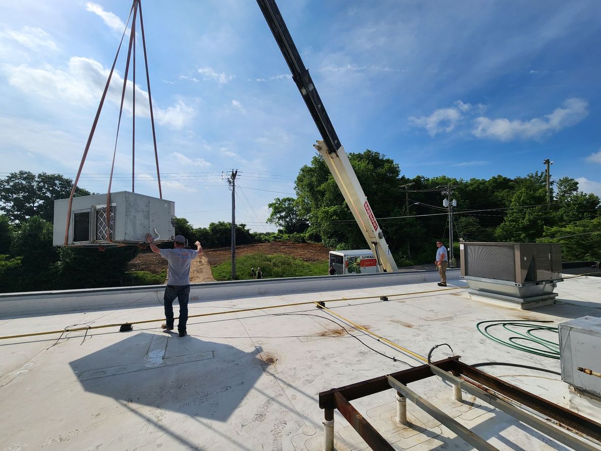A crane lifting a large HVAC unit onto a rooftop, with workers supervising under a sunny sky.