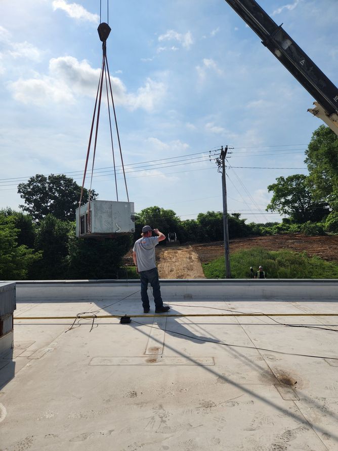 A man on a rooftop watches a crane lift a large white box. Trees and power lines in the background.