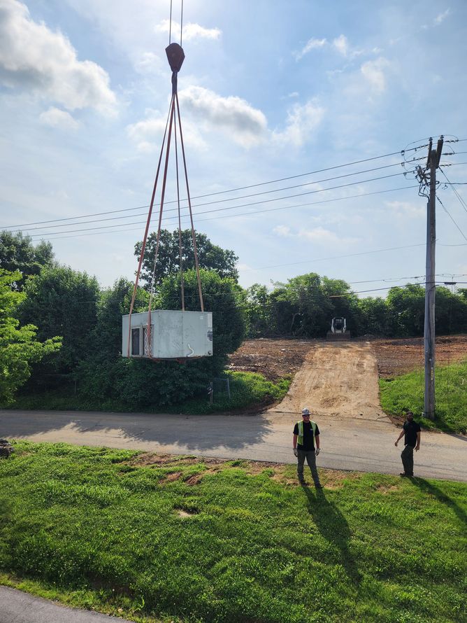 A crane lifts a large, light-colored rectangular object over green foliage and a dirt road; two people watch.