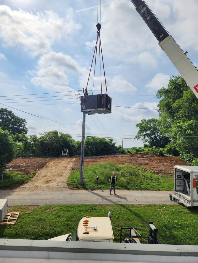 A crane lifts a large, gray transformer above a utility pole, surrounded by grass, trees, and a blue sky.