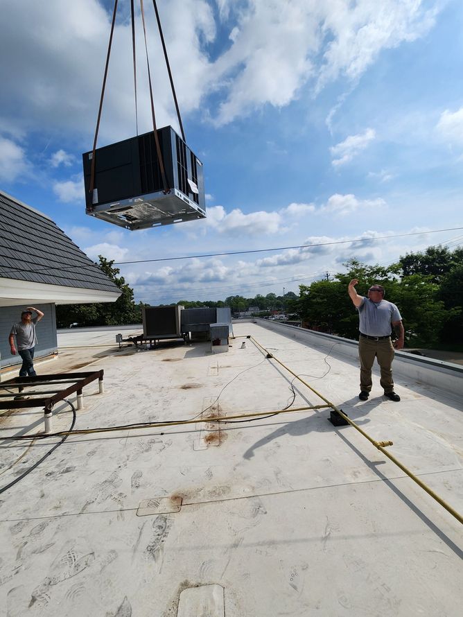 Workers installing HVAC unit on a rooftop, unit suspended from a crane, sunny day.