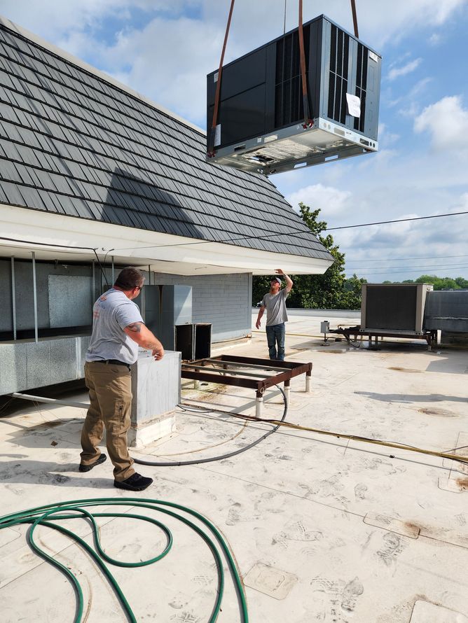 Two men on a rooftop installing an HVAC unit; a crane lifts a large metal air conditioner.