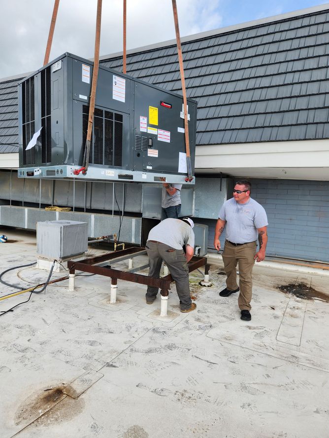Workers installing an HVAC unit on a rooftop. A crane lifts the unit above a platform, while workers secure it.
