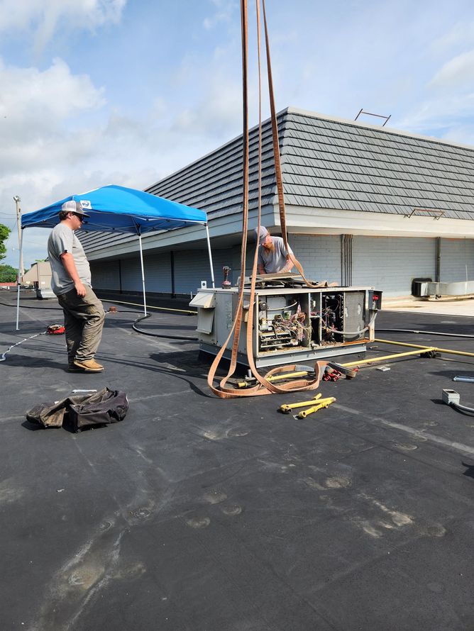 Two workers replacing HVAC unit on a building's flat roof. Brown straps lift the unit.