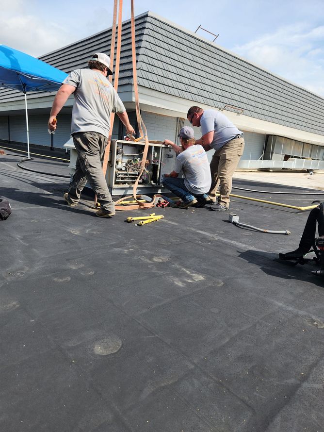 Three workers installing rooftop HVAC unit on a dark asphalt surface. Blue tent in the background.