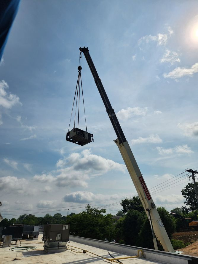 A crane lifting an HVAC unit onto a commercial building's roof under a blue sky.