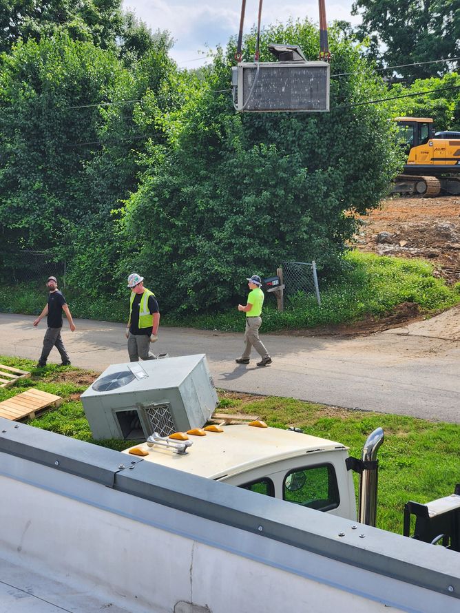 Workers move HVAC unit with crane near a truck and construction site, with greenery in background.