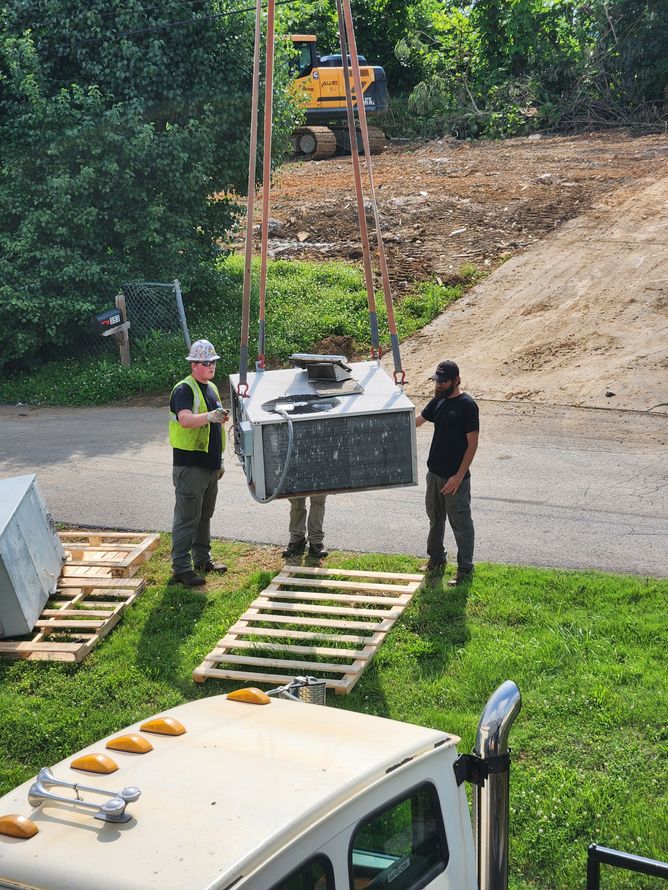 Two workers lift a large metal unit with straps near a truck; a backhoe works in the background.