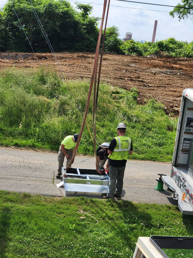 Three utility workers lifting equipment with a crane on a road, near a grassy area and trees.