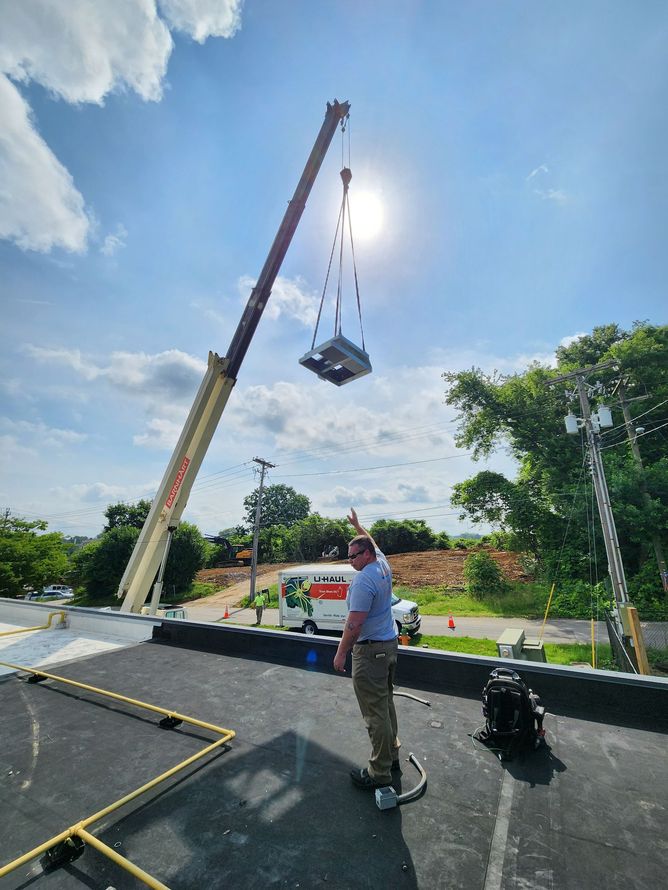 A crane lifts an HVAC unit onto a rooftop with a worker watching under a sunny sky.