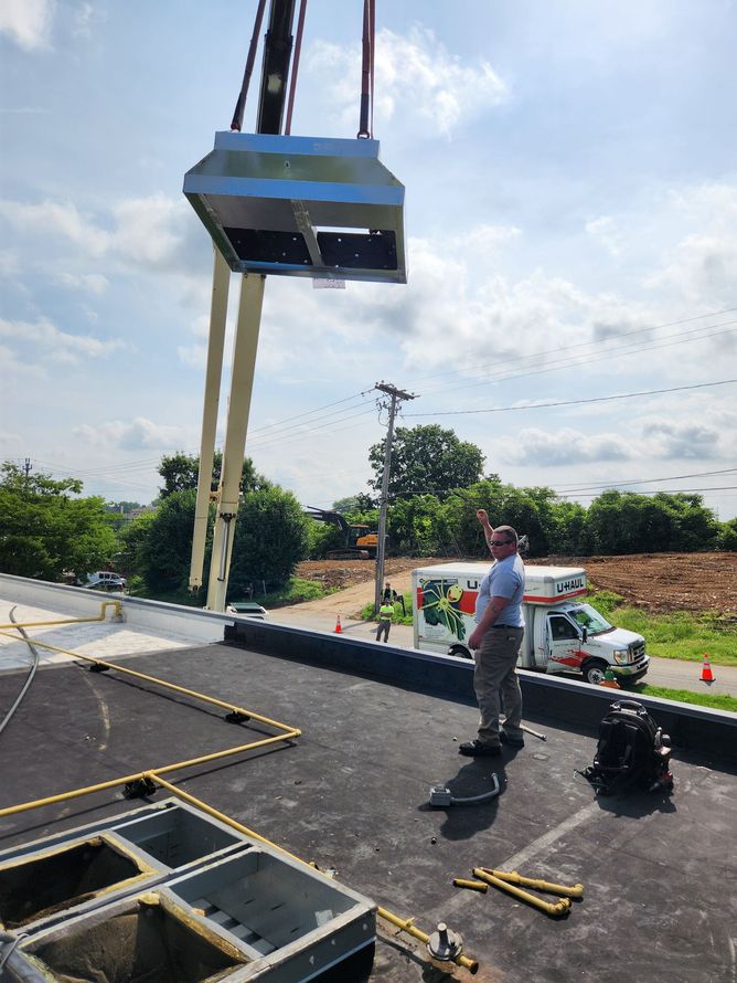 Crane lifting HVAC unit onto a rooftop; worker watches, with ambulance and trees visible in the background.