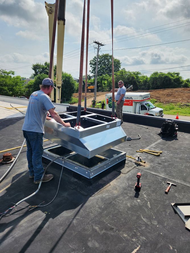 Two men installing a rooftop vent. Metal vent suspended by crane cables, on a black tar surface.