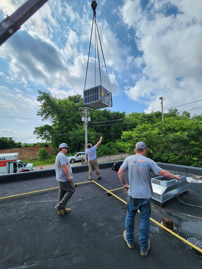 Workers installing HVAC unit on a roof. Crane lifting unit, workers guiding it. Blue sky, trees in background.
