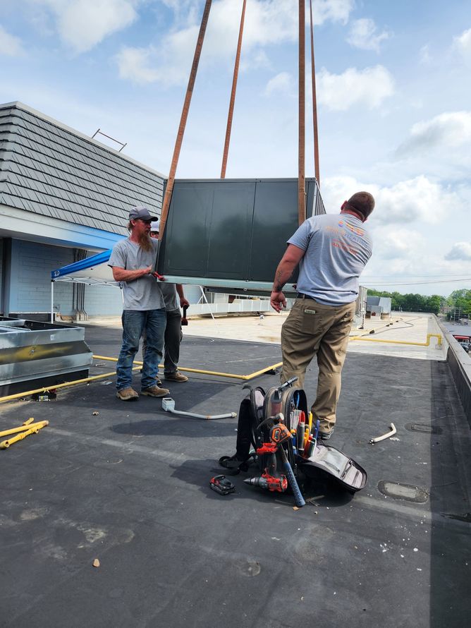 Three workers lift an HVAC unit on a rooftop. The unit is suspended by cranes. Tools are visible.