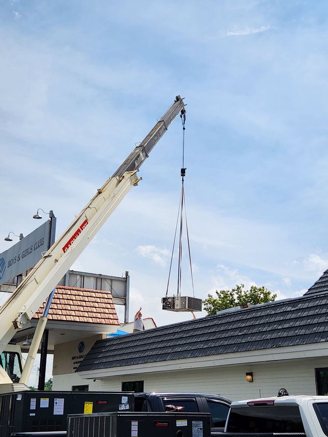 A crane lifting a rectangular object near a building with a dark roof under a cloudy sky.