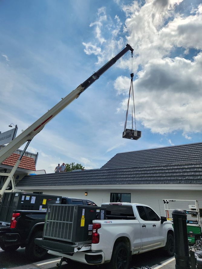 A crane lifts equipment over a house. White trucks and other gear are visible below a cloudy sky.