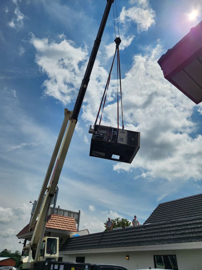 A crane lifts a large, dark unit toward a rooftop under a cloudy blue sky.