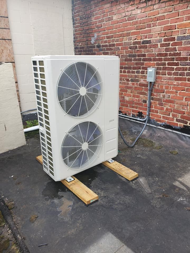 A large white air conditioner is sitting on wooden stands in front of a brick building.