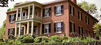 A large famous brick house with a porch and shutters is surrounded by trees and bushes in Church Hill Tennessee