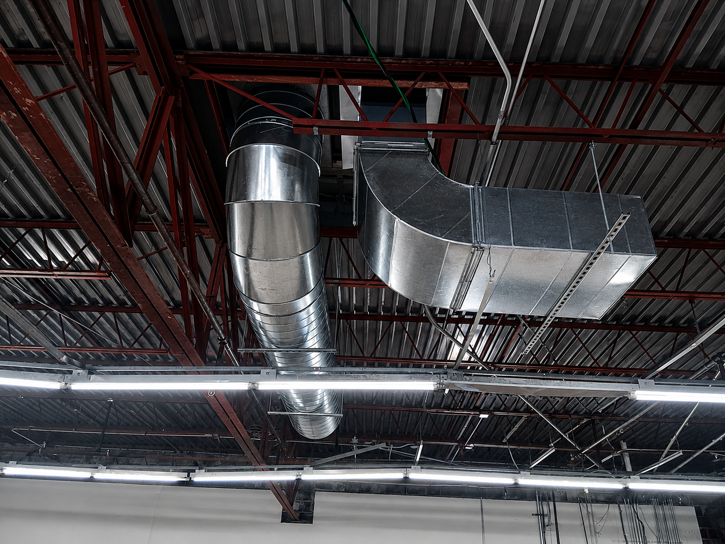 Industrial ceiling with silver HVAC ducts and fluorescent lights against a red metal roof frame