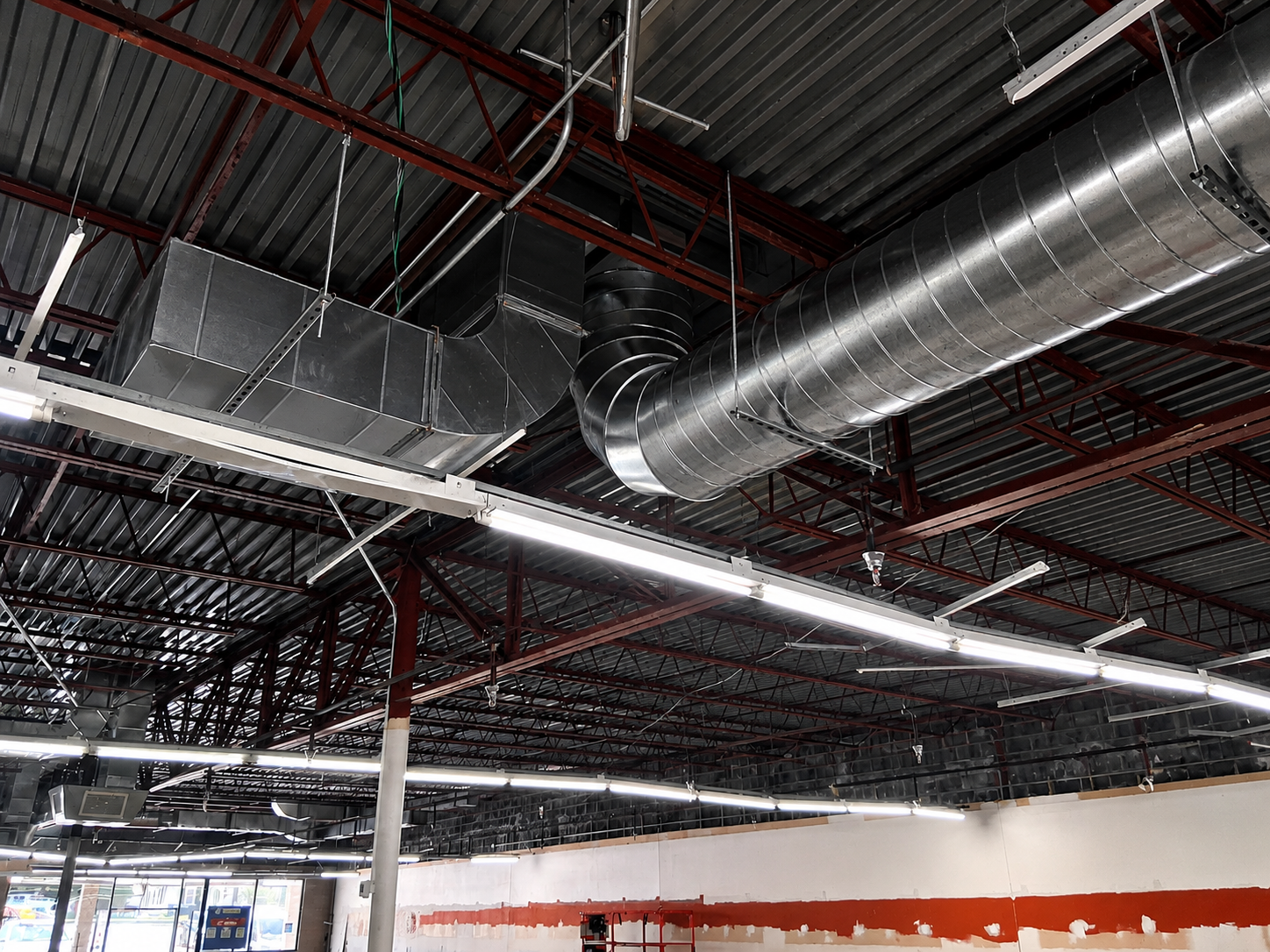 Industrial warehouse ceiling with exposed metal ducts, red beams, and fluorescent lights