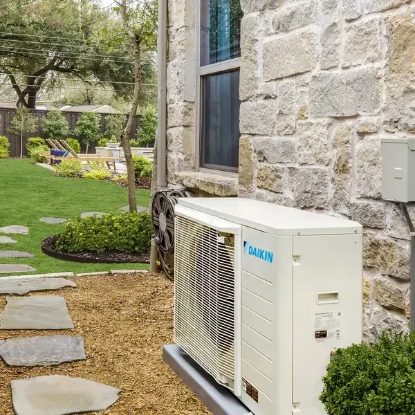 A beige Daikin outdoor HVAC unit sits on a stone patio next to a stone-walled house, with a grassy yard in the background.