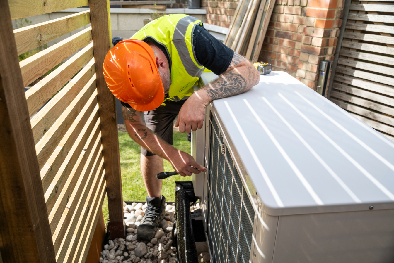 HVAC technician in orange hard hat and safety vest repairs an air conditioning unit outdoors.