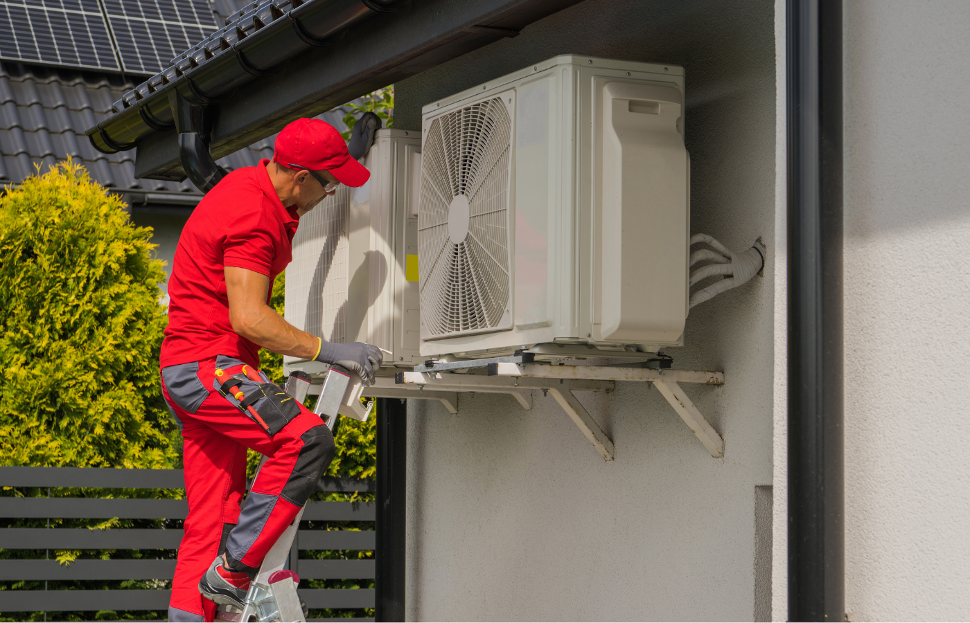 A technician in red jumpsuit, repairing an air conditioning unit, mounted on a white wall, outside a house.