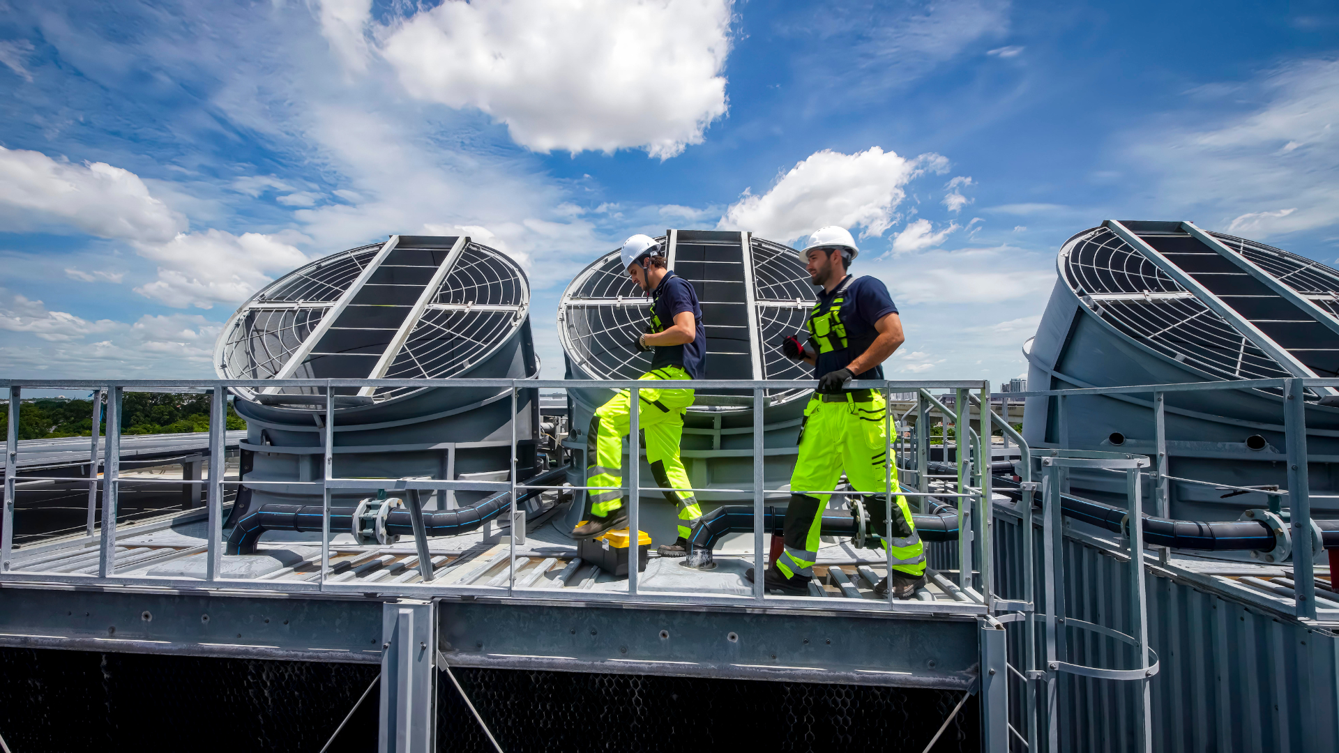 Two technicians in safety gear and hard hats work on large industrial cooling fans atop a rooftop under a blue sky.