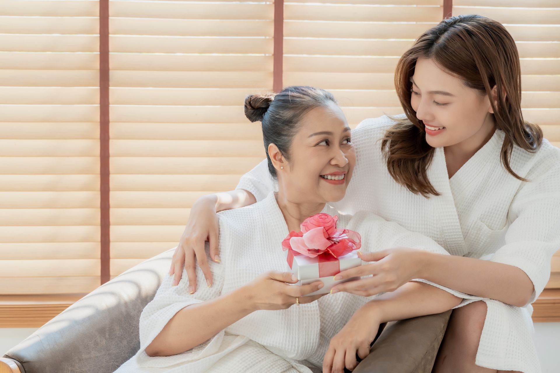 Two women smiling on a couch, sharing a small pink gift in a bright living room.