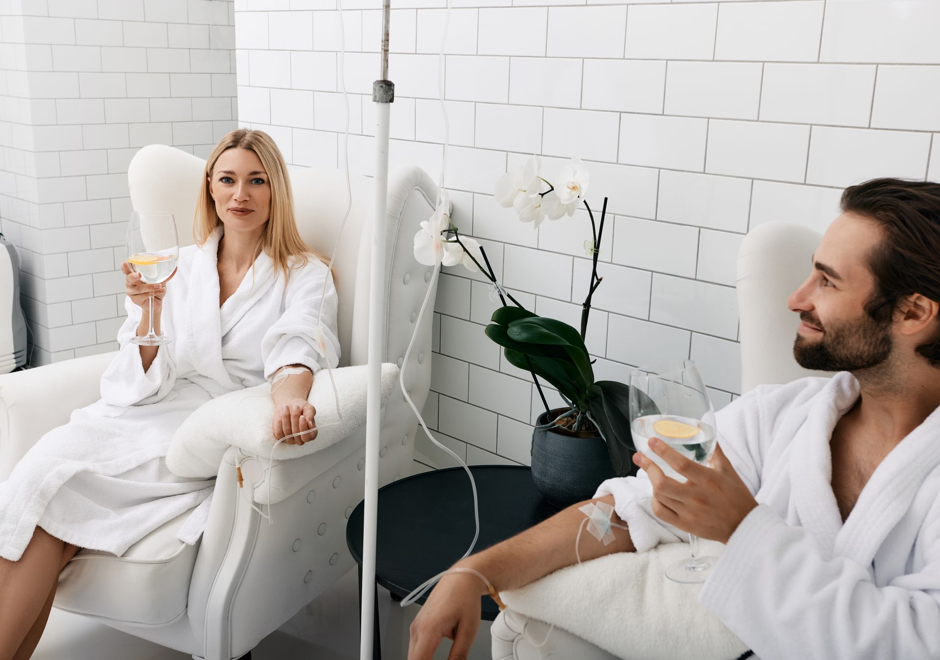Two people in white robes relax in chairs, holding drinks in a bright spa-like room.